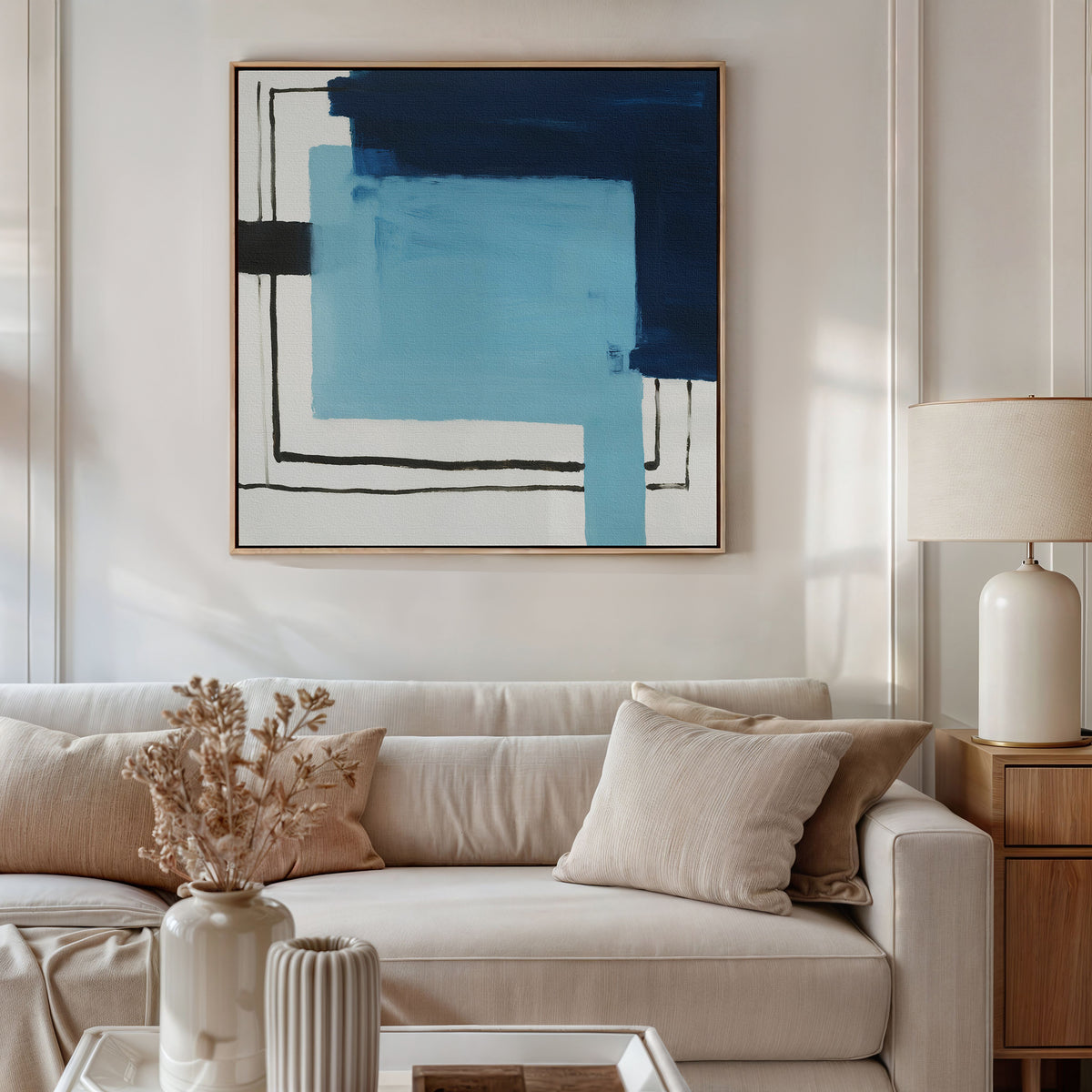 A modern living room with a beige sofa, light cushions, a side table with a cream lamp, and the striking Cobalt Intersection Canvas Art - Square on the wall. A vase with dried flowers and a ribbed ceramic decor piece grace the coffee table.
