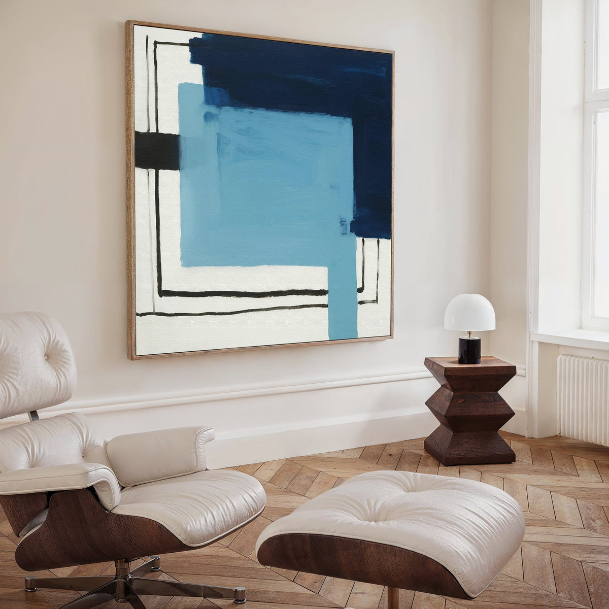 A modern living room with a white leather lounge chair, geometric wooden side table, small white lamp, and the striking Cobalt Intersection Canvas Art - Square in blue, navy, and black tones as contemporary wall art.