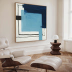 A modern living room with a white leather lounge chair, geometric wooden side table, small white lamp, and the striking Cobalt Intersection Canvas Art - Square in blue, navy, and black tones as contemporary wall art.
