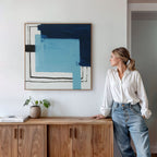 A woman in a white blouse and jeans leans against a wooden cabinet, gazing at the Cobalt Intersection Canvas Art - Square with blue and black geometric shapes on a white wall. A small plant and books rest on the cabinet.