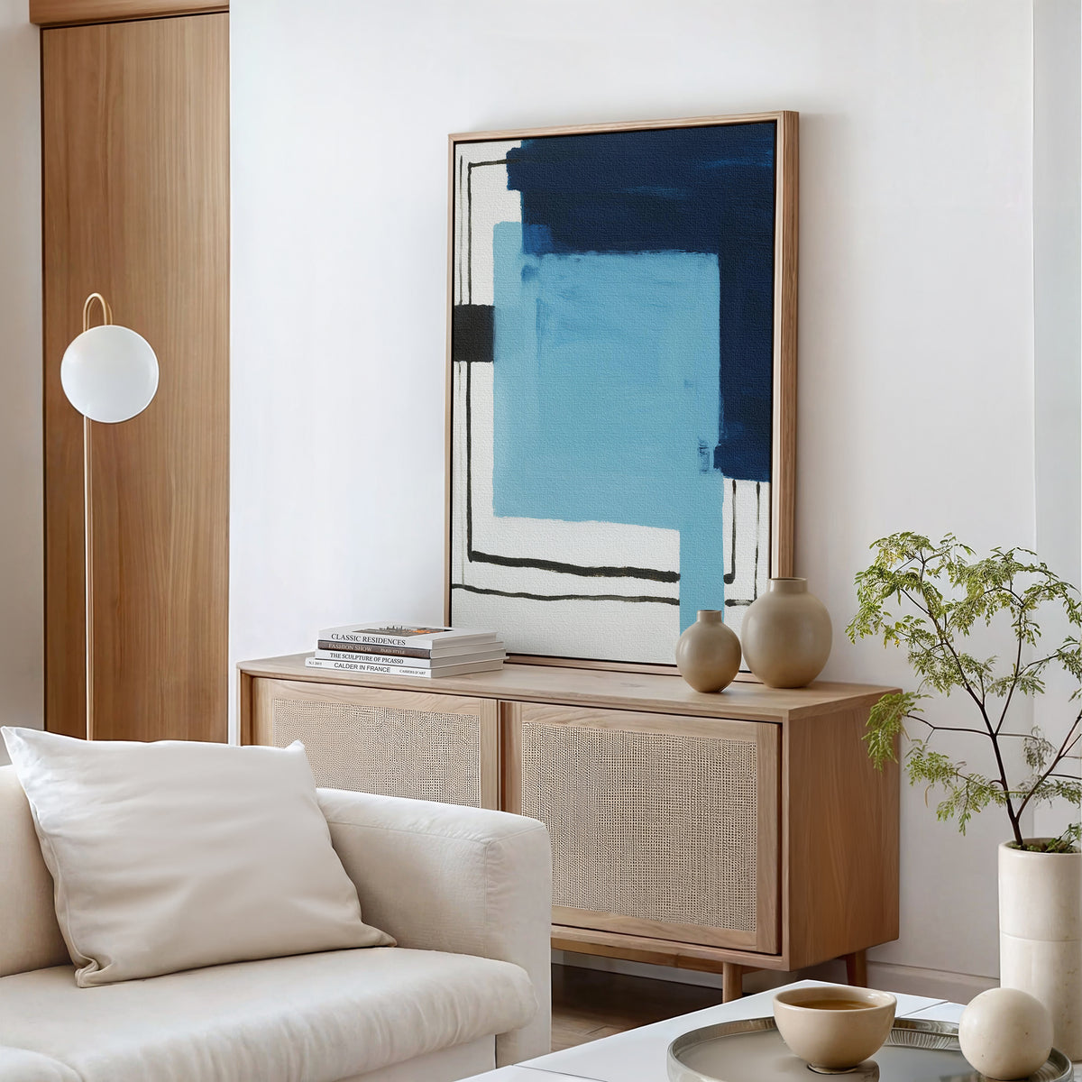A modern living room featuring the Cobalt Intersection Canvas Art - Vertical, a light wood sideboard, potted plant, white sofa, round coffee table with ceramics, and globe floor lamp—ideal for fans of modern home decor.