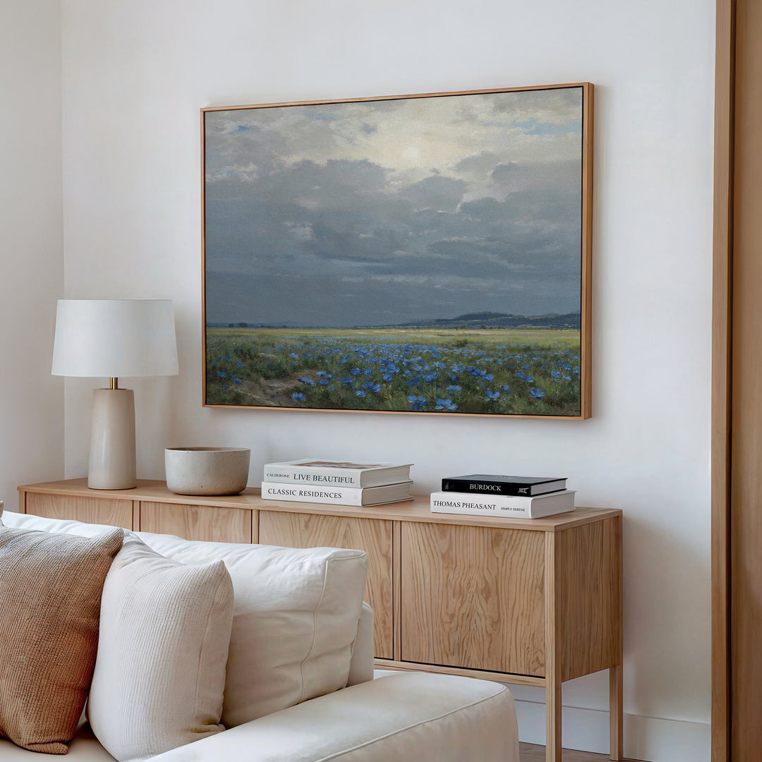 A modern living room with a light wood sideboard, white table lamp, stacked books, and the "Cornflower Fields Canvas Art - Horizontal," featuring a cloudy sky over blue wildflowers—perfect for countryside home decor.