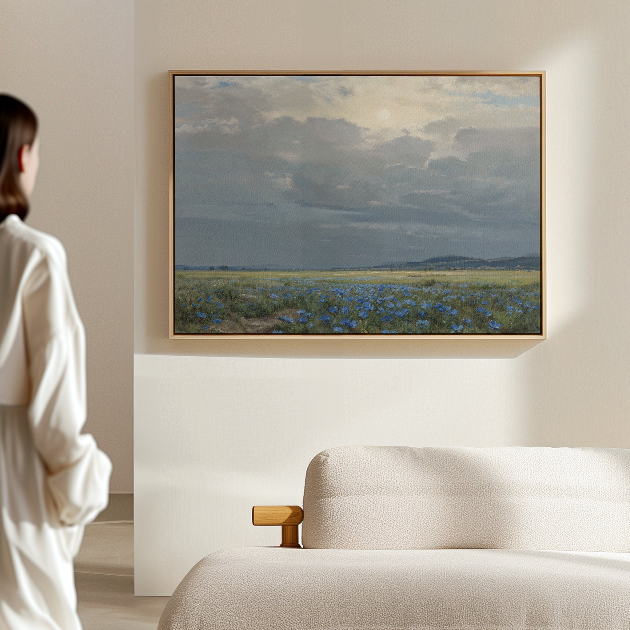 A person in a white shirt admires the Cornflower Fields Canvas Art - Horizontal, a pastoral landscape with blue blooms and cloudy sky, hanging above a cream-colored sofa in a minimal, light-filled room.