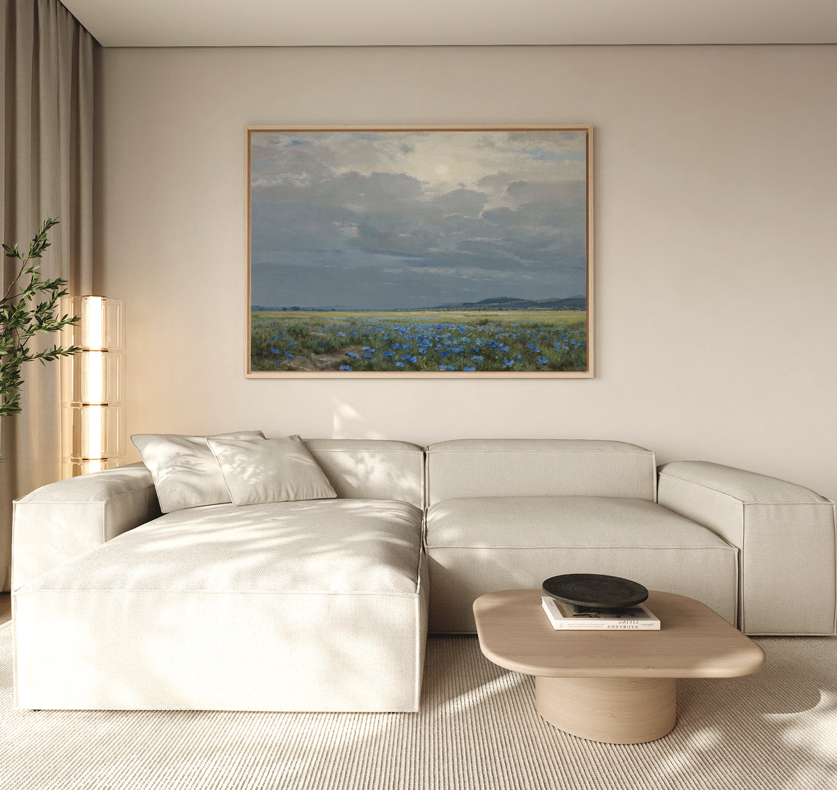 A modern living room with a cream sectional, light wood coffee table, potted plant, and the Cornflower Fields Canvas Art - Horizontal featuring blue flowers and a cloudy sky above the sofa—perfect for countryside decor.