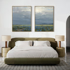 A modern bedroom features an olive green bed, white bedding, round nightstands with beige lamps, and above the bed hangs Cornflower Fields - Set of 2: large canvas prints of pastoral landscapes with cloudy skies and cornflower fields.