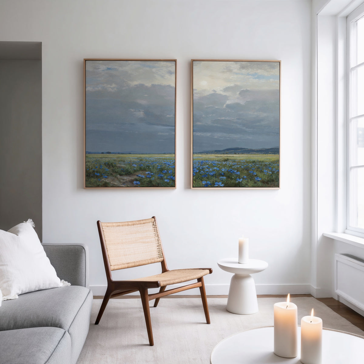 A modern living room features a wooden chair, round white side table, lit candles, and natural light. On the white wall above hang the "Cornflower Fields - Set of 2" prints, showing cloudy skies and blue flowers in cornflower fields.
