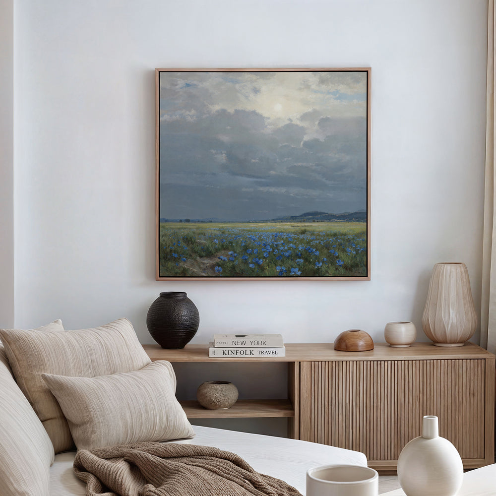 A modern, neutral-toned living room showcases a wooden sideboard with decor, and above it hangs the Cornflower Fields Canvas Art - Square, featuring a cloudy sky over cornflower fields on the white wall.