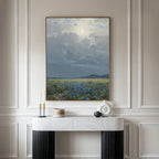 A marble console table with minimalist decor stands against a wall, topped by the Cornflower Fields Canvas Art - Vertical, which features a pastoral landscape of cloudy skies over cornflower fields and distant hills.