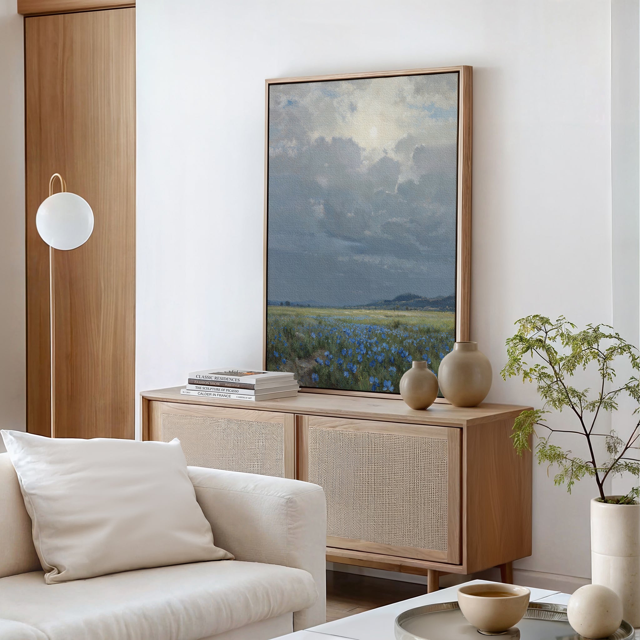 A modern living room with a light wood sideboard, pottery vases, a vertical Cornflower Fields Canvas Art, a small green plant, and a white sofa with neutral pillows. The minimal decor creates a calming atmosphere.