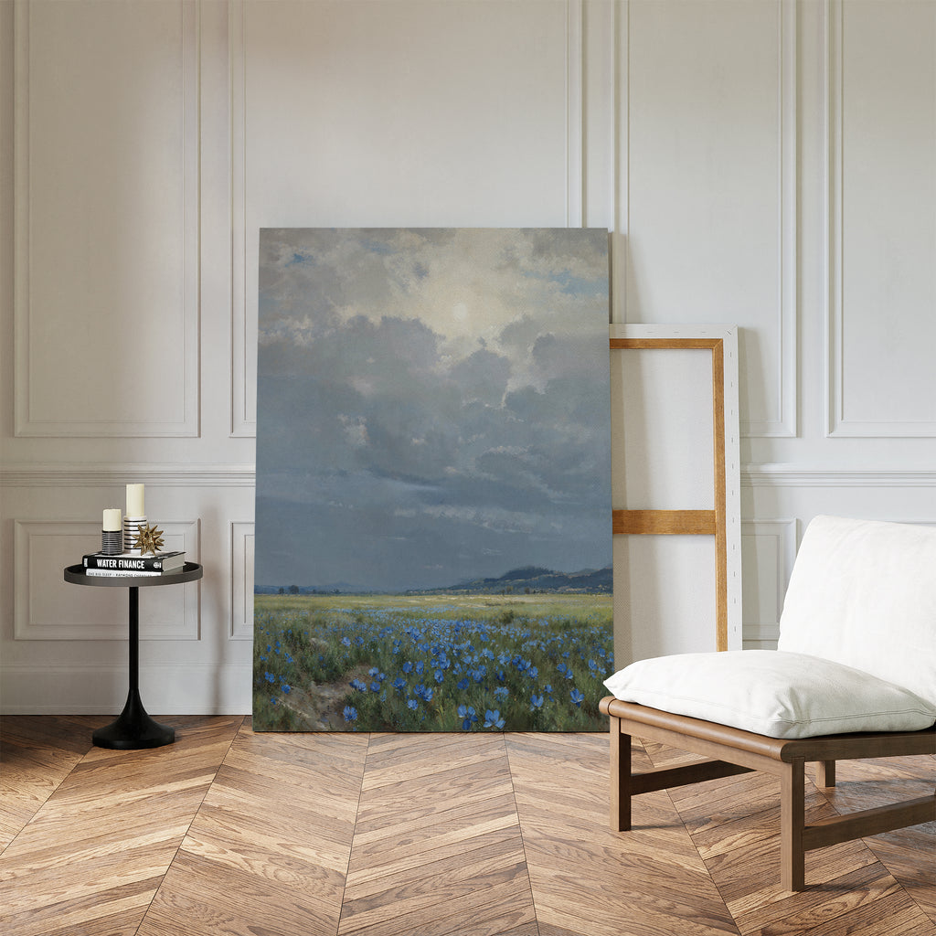 The Cornflower Fields Canvas Art - Vertical leans against a white paneled wall in a stylish room featuring a wood floor, white cushioned chair, and a small black table decorated with candles and books.