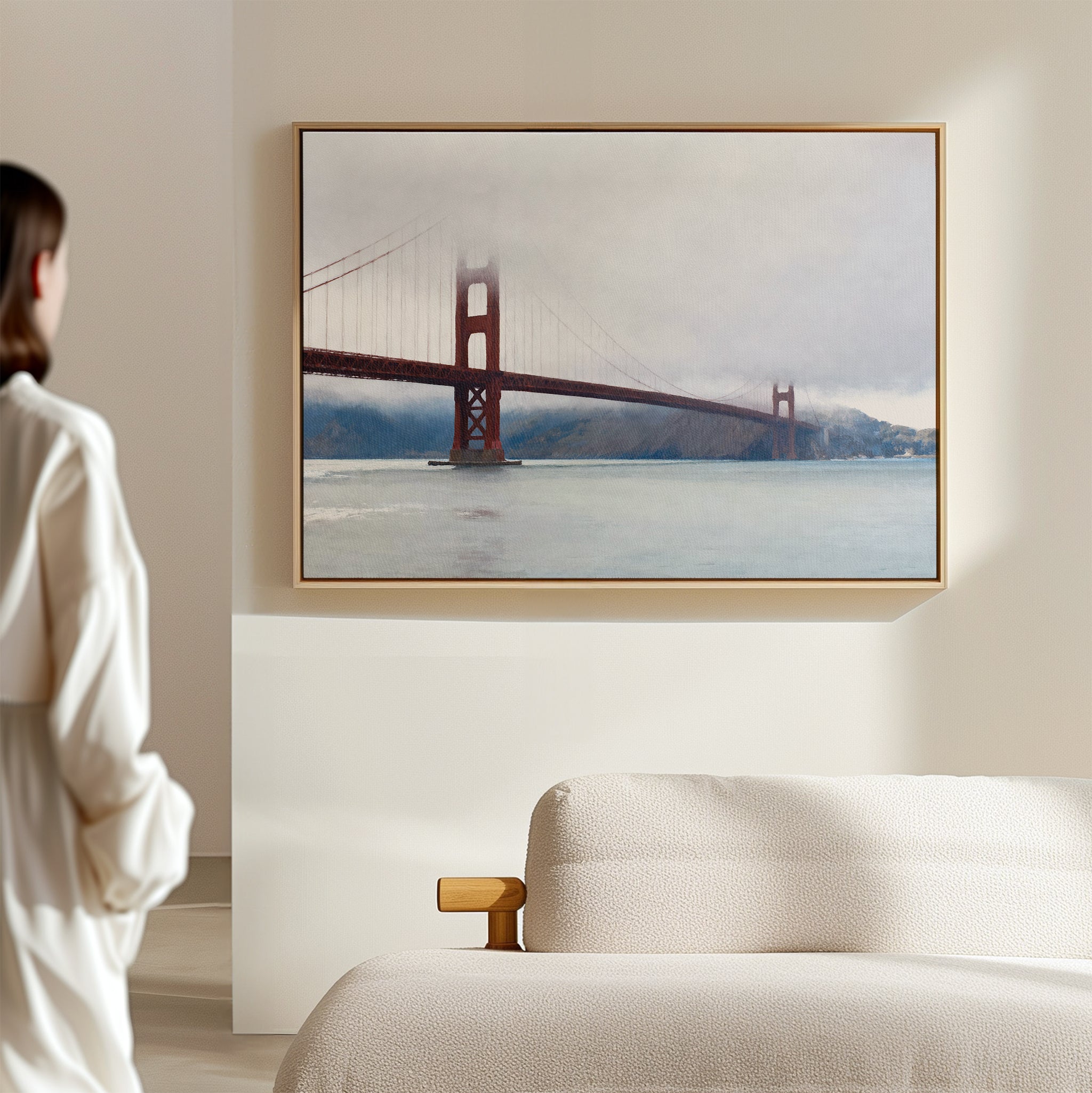 A woman admires the Crimson Passage Canvas Art - Horizontal, featuring the Golden Gate Bridge in fog, as it hangs above a cream-colored sofa in a minimal, light-filled room, adding a touch of coastal elegance.