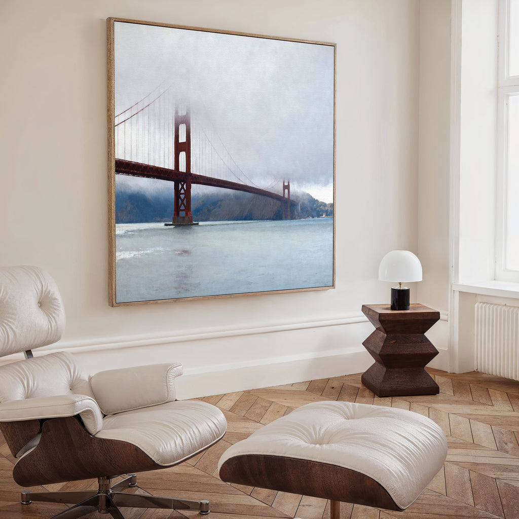 A modern, minimalist living room with a white lounge chair, ottoman, wooden side table, lamp, and the Crimson Passage Canvas Art - Square showcasing the Golden Gate Bridge in fog for a touch of coastal elegance.