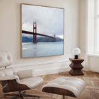 A modern, minimalist living room with a white lounge chair, ottoman, wooden side table, lamp, and the Crimson Passage Canvas Art - Square showcasing the Golden Gate Bridge in fog for a touch of coastal elegance.
