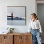 A woman in a white shirt and jeans leans on a wooden sideboard, admiring the Crimson Passage Canvas Art - Square. A coastal touch is added by the nearby potted plant and book.