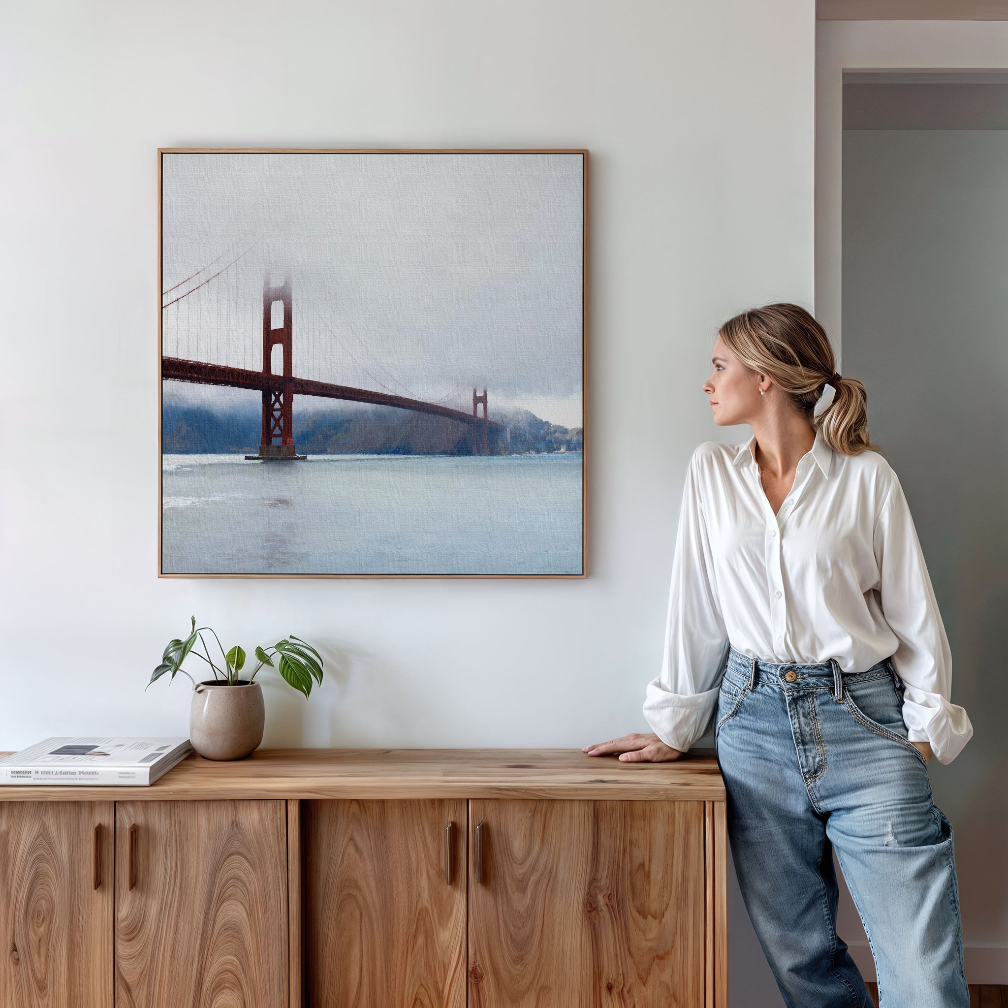 A woman in a white shirt and jeans leans on a wooden sideboard, admiring the Crimson Passage Canvas Art - Square. A coastal touch is added by the nearby potted plant and book.