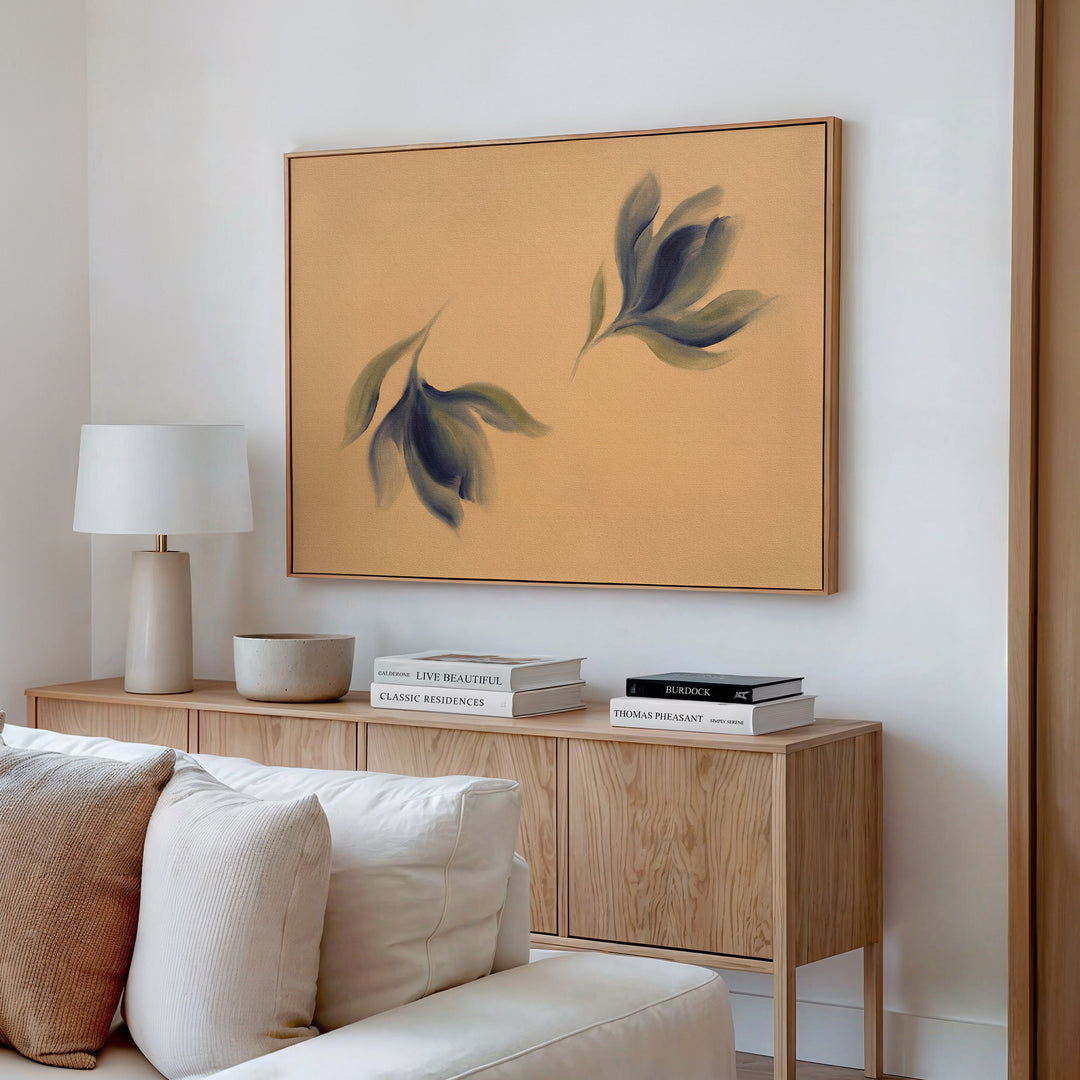 A modern living room corner features a light wood sideboard, white table lamp, bowl, stacked books, beige sofa, and above it, the Dancing Leaves Canvas Art - Horizontal showcasing abstract blue-green leaves as bold botanical wall decor.