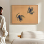 A person in white stands in a modern minimal living room, admiring the Dancing Leaves Canvas Art - Horizontal, featuring soft blue and green leaf shapes on muted beige, displayed above a textured cream sofa.