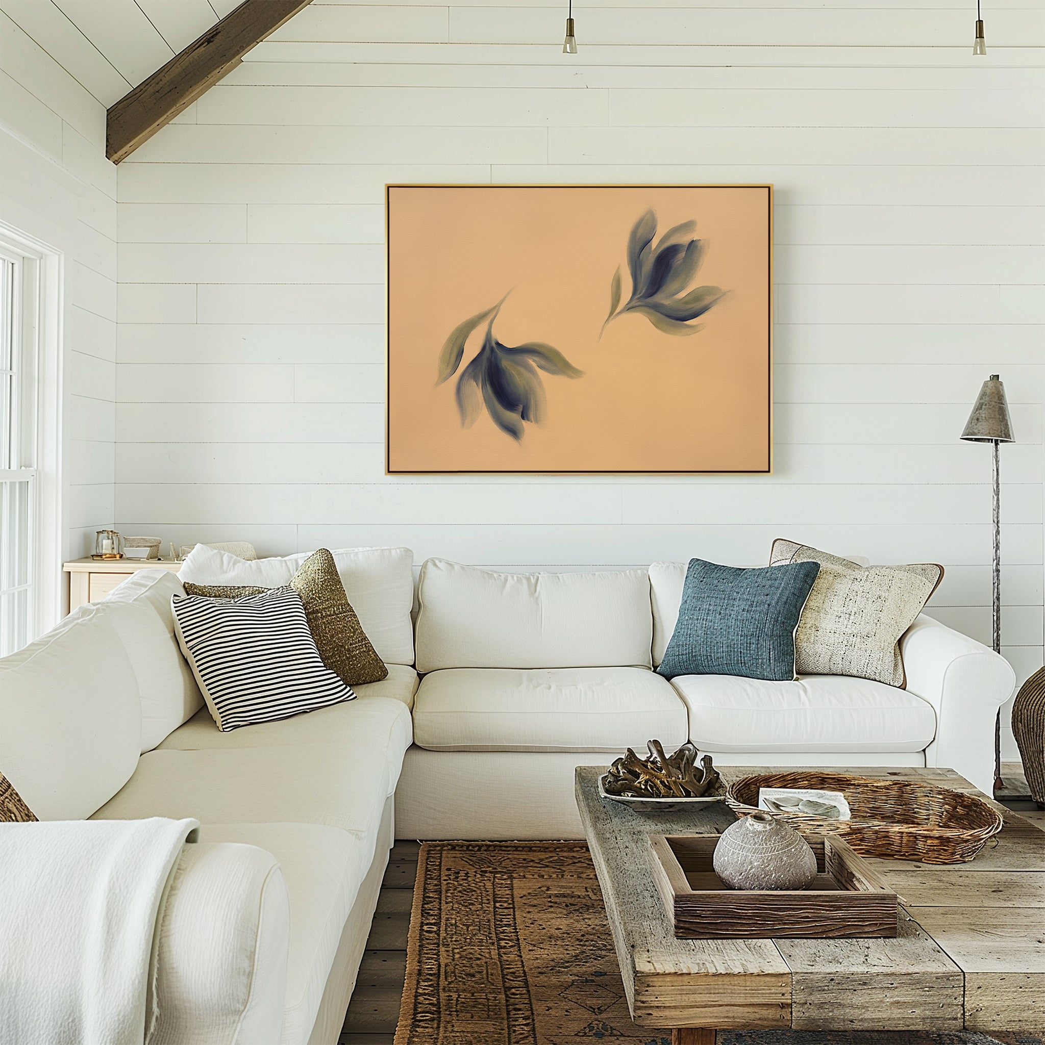 A cozy living room with a white sectional sofa, assorted pillows, a rustic wooden coffee table, and the Dancing Leaves Canvas Art - Horizontal—a large framed print of abstract leaves on a peach background—adorning the white shiplap wall.