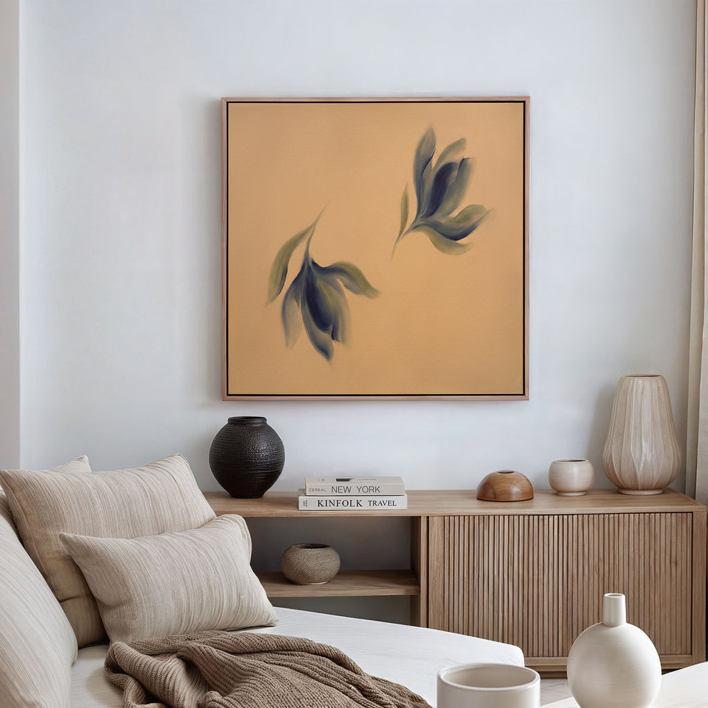 A minimalist living room with a beige sofa, neutral cushions, wooden sideboard, ceramic vases, books, and the Dancing Leaves Canvas Art - Square featuring green and blue leaves on a tan background displayed on the wall.