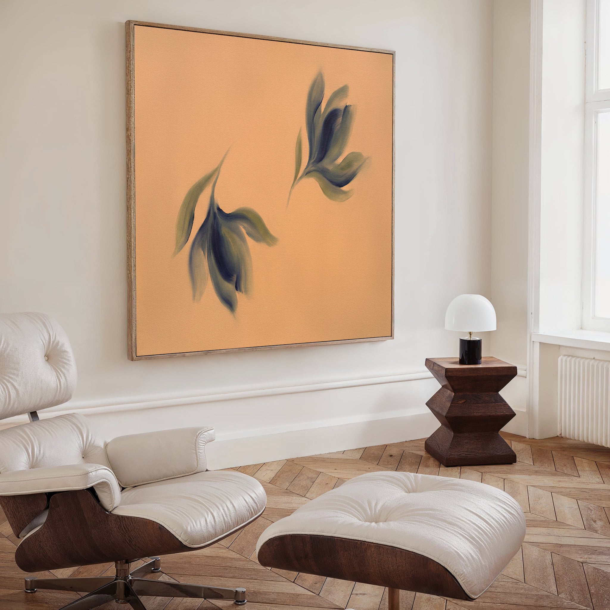 A modern living room features a white leather lounge chair with ottoman, a dark wood side table with a white lamp, and Nordic Park vibes thanks to the Dancing Leaves Canvas Art - Square—a bold blue and green leaf print on peach.