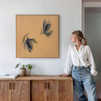 A woman in a white blouse and jeans stands by a wooden cabinet, admiring the Dancing Leaves Canvas Art - Square, which features abstract leaves on a tan background. A potted plant and books are placed on the cabinet beneath the artwork.