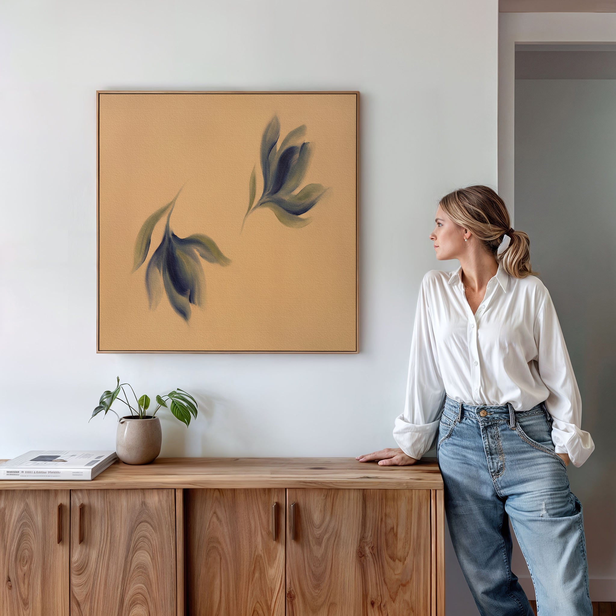 A woman in a white blouse and jeans stands by a wooden cabinet, admiring the Dancing Leaves Canvas Art - Square, which features abstract leaves on a tan background. A potted plant and books are placed on the cabinet beneath the artwork.