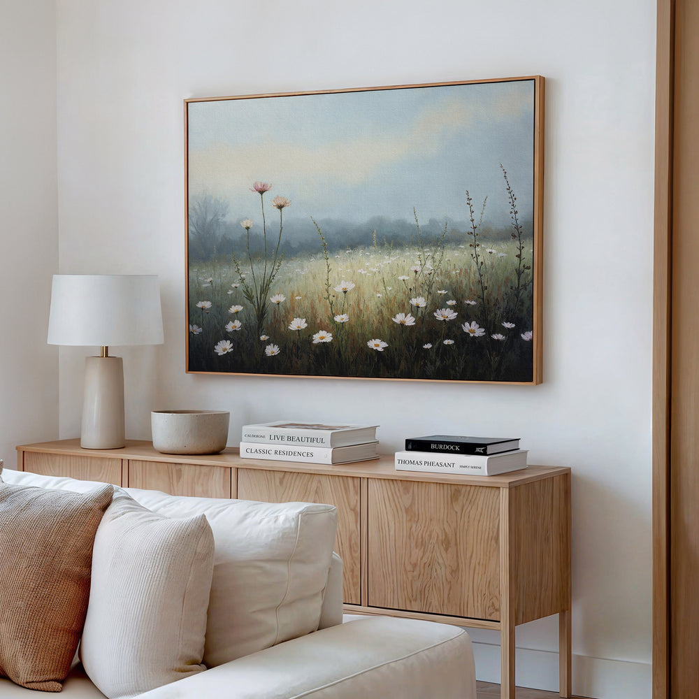 A cozy living room corner features a light wood sideboard with books, a lamp, and a bowl. Above, the Dawn Meadow Canvas Art - Horizontal adds botanical charm to the white wall. A cream-colored sofa is in the foreground.
