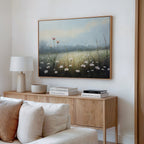 A cozy living room corner features a light wood sideboard with books, a lamp, and a bowl. Above, the Dawn Meadow Canvas Art - Horizontal adds botanical charm to the white wall. A cream-colored sofa is in the foreground.