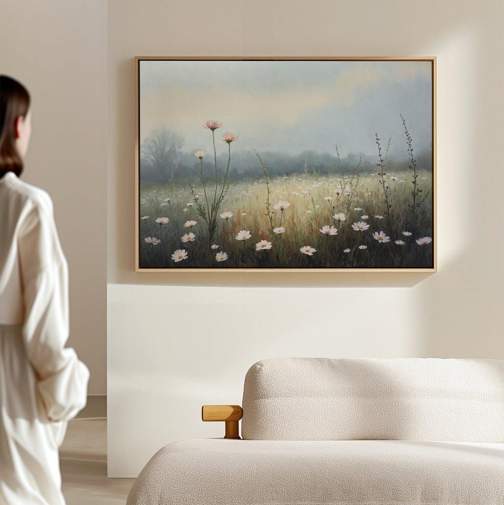 A person in a white robe admires the Dawn Meadow Canvas Art—a framed wildflower print hanging above a beige couch in a softly lit, minimalistic room.