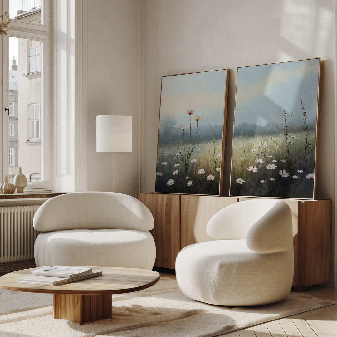 A modern living room with two curved white armchairs, a round wooden coffee table, a floor lamp, and the Dawn Meadow - Set of 2 wildflower field art on a wooden cabinet. Sunlight streams through tall windows, enhancing the serene decor.