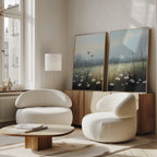 A modern living room with two curved white armchairs, a round wooden coffee table, a floor lamp, and the Dawn Meadow - Set of 2 wildflower field art on a wooden cabinet. Sunlight streams through tall windows, enhancing the serene decor.