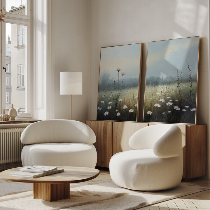 A modern living room with two curved white armchairs, a round wooden coffee table, a floor lamp, and the Dawn Meadow - Set of 2 wildflower field art on a wooden cabinet. Sunlight streams through tall windows, enhancing the serene decor.
