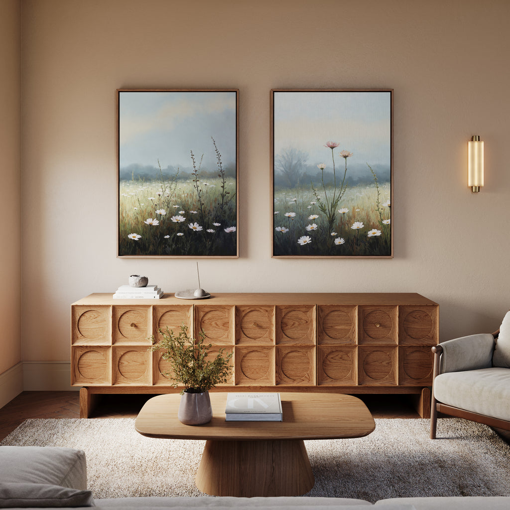 A cozy living room features a wooden sideboard with a geometric pattern, a matching coffee table, a white armchair, and the “Dawn Meadow - Set of 2” wildflower art above the sideboard. A wall sconce provides soft lighting.