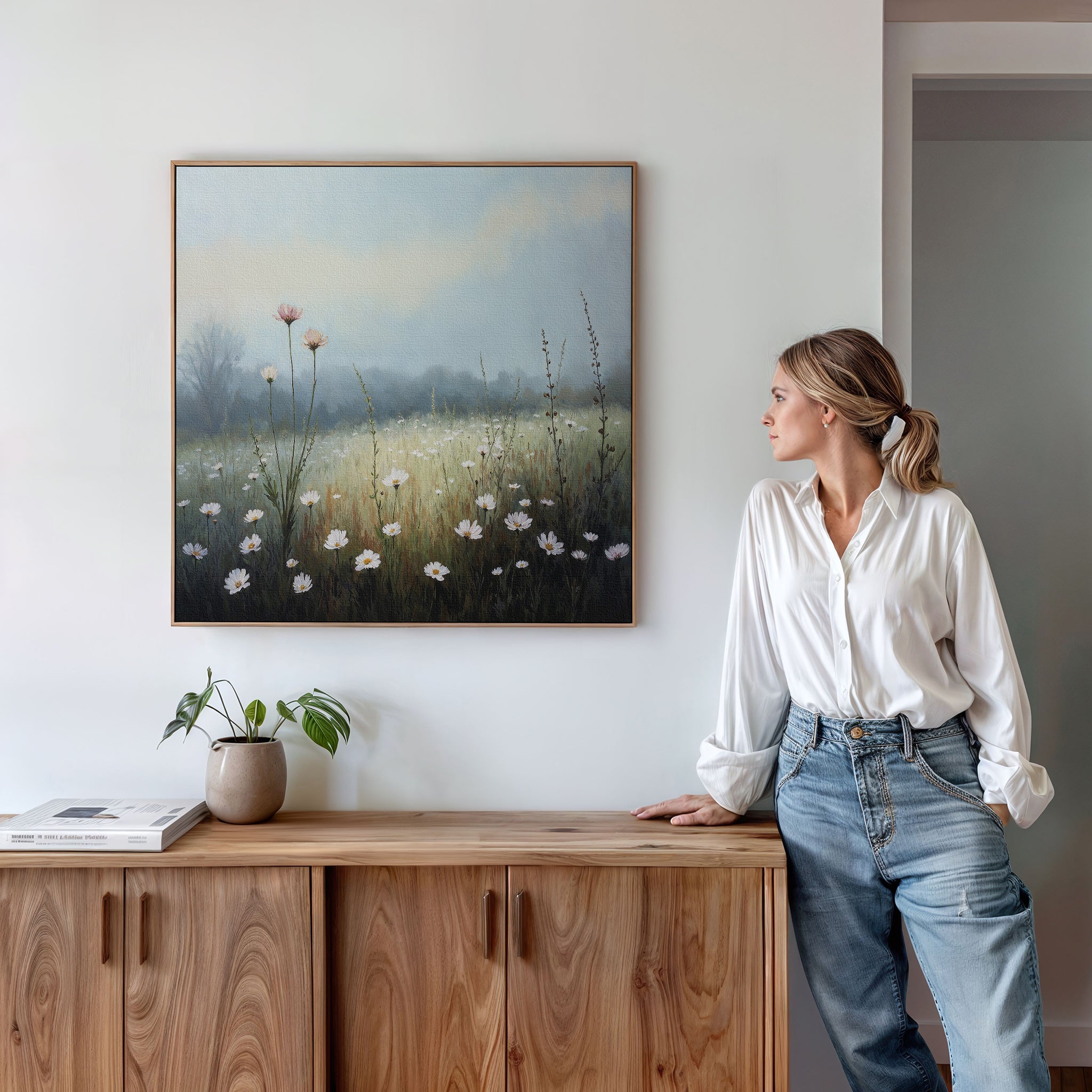 A woman in a white blouse and jeans leans against a wooden cabinet, gazing at the Dawn Meadow Canvas Art - Square. A small plant and books on the cabinet complete the serene bedroom decorated with this botanical wall art.