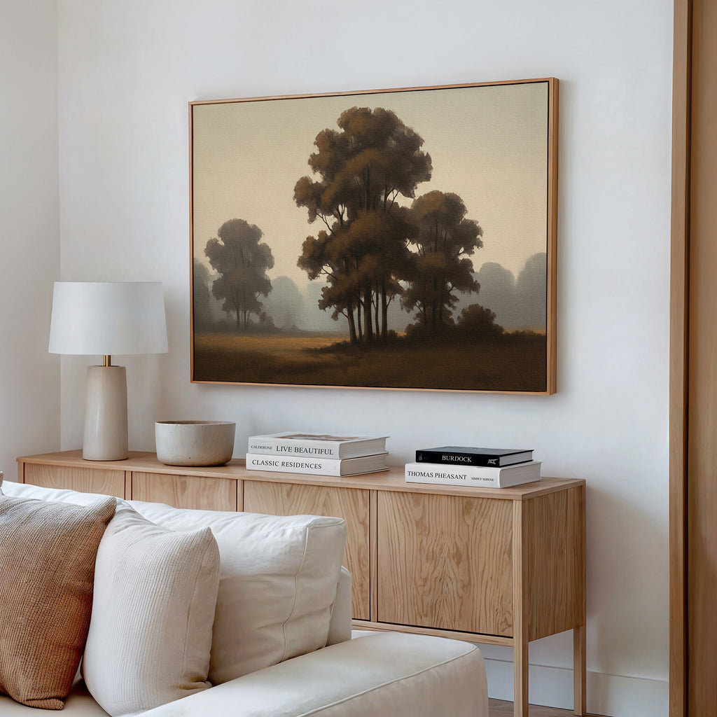 A cozy living room features a wooden sideboard with books and decor, a white table lamp, and the "Dawn Silhouettes Canvas Art - Horizontal" in soft brown tones above a pale sofa.