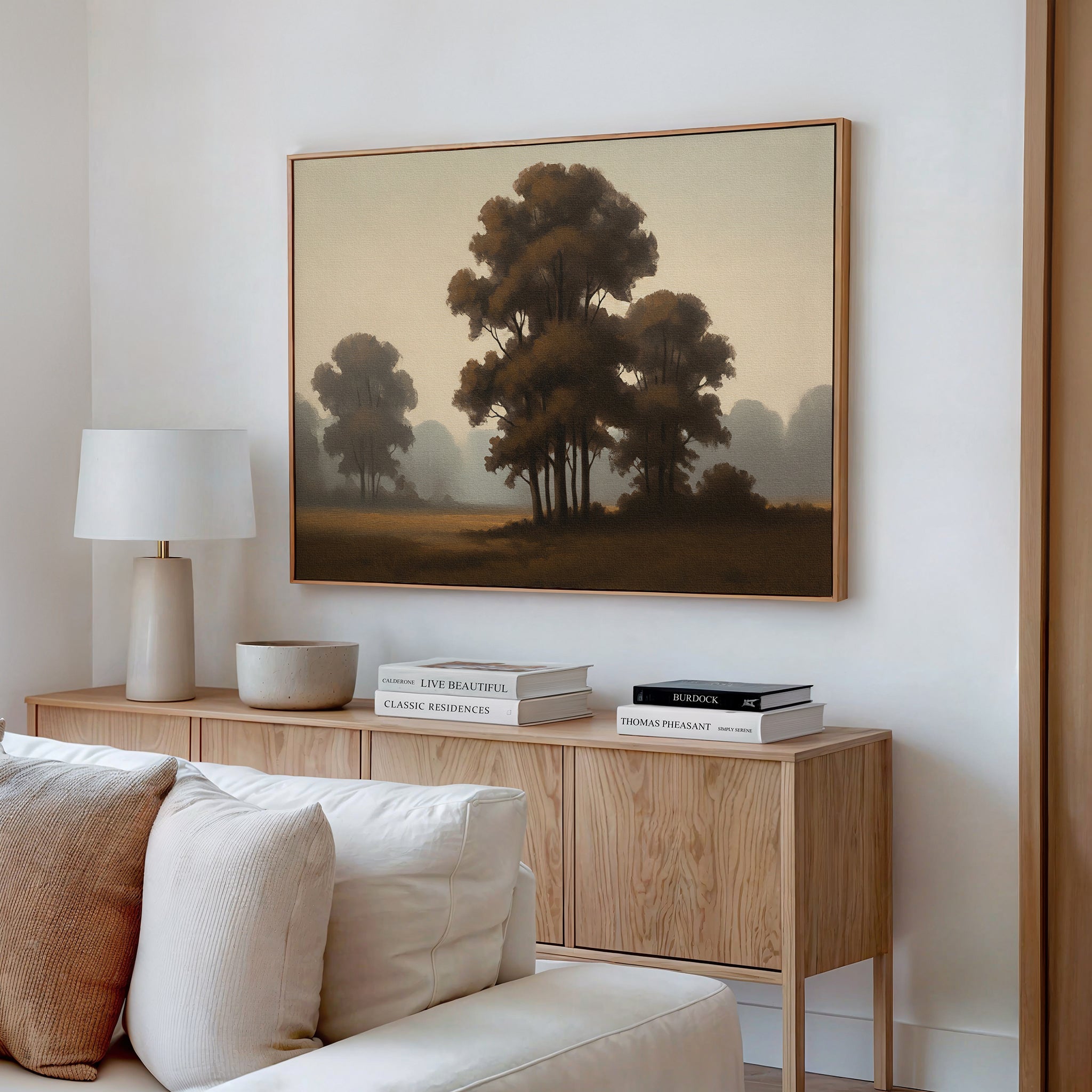 A cozy living room features a wooden sideboard with books and decor, a white table lamp, and the "Dawn Silhouettes Canvas Art - Horizontal" in soft brown tones above a pale sofa.