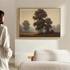 A person in white stands in a modern room with a beige sofa, admiring the "Dawn Silhouettes Canvas Art - Horizontal," which features tall trees and a misty field on the wall, adding calm Nordic Park beauty to the space.