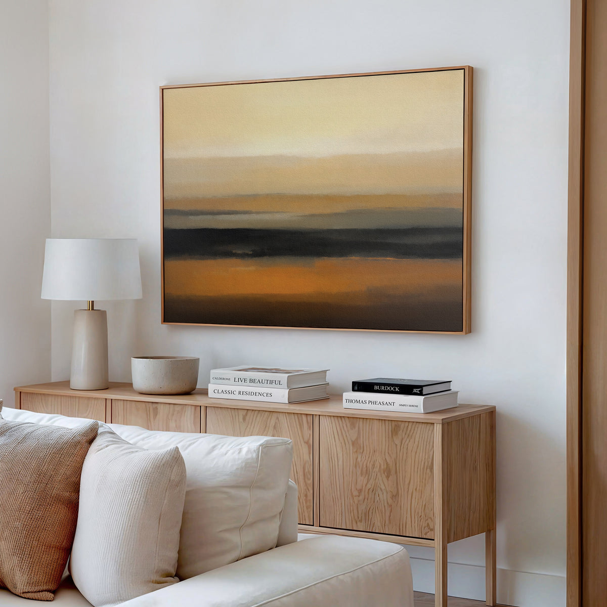 A modern living room features a wooden sideboard, white lamp, stacked books, and a bowl. Above the light sofa with textured pillows hangs the Desert Mirage Canvas Art - Horizontal in earth tones, adding warmth to the space.