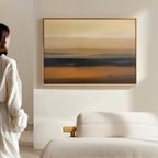 A woman in a white blouse stands in a bright, minimal room, admiring the Desert Mirage Canvas Art - Horizontal with warm earth tones and dark stripes, displayed above a cream-colored sofa.