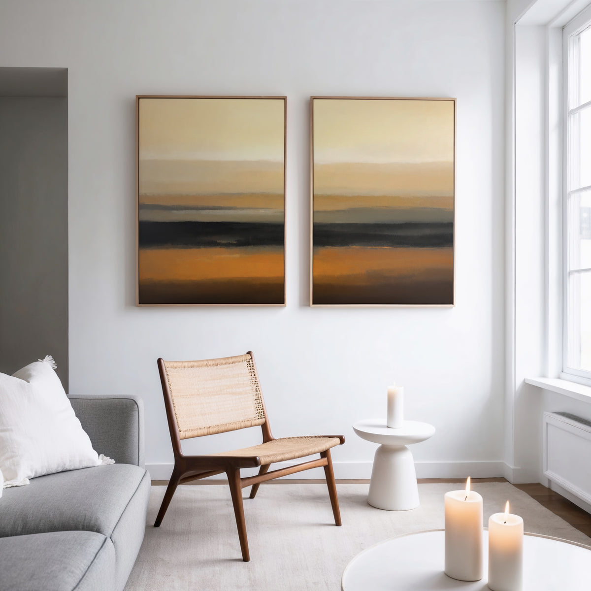 A modern living room with a light gray sofa, woven chair, small white side table, and three lit candles. The Desert Mirage - Set Of 2 abstract prints with warm earth tones hang above the chair. Large windows fill the space with light.