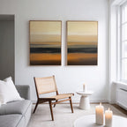 A modern living room with a light gray sofa, woven chair, small white side table, and three lit candles. The Desert Mirage - Set Of 2 abstract prints with warm earth tones hang above the chair. Large windows fill the space with light.