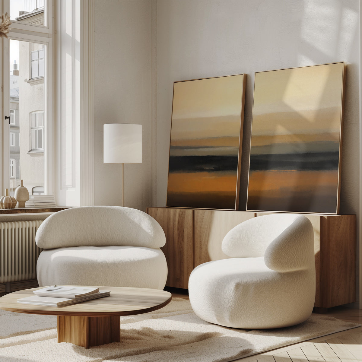 A modern living room with two white, rounded armchairs, a low wooden coffee table, light wood sideboard, and "Desert Mirage - Set Of 2" abstract desert landscape art in earth tones. Sunlight pours in through large windows.