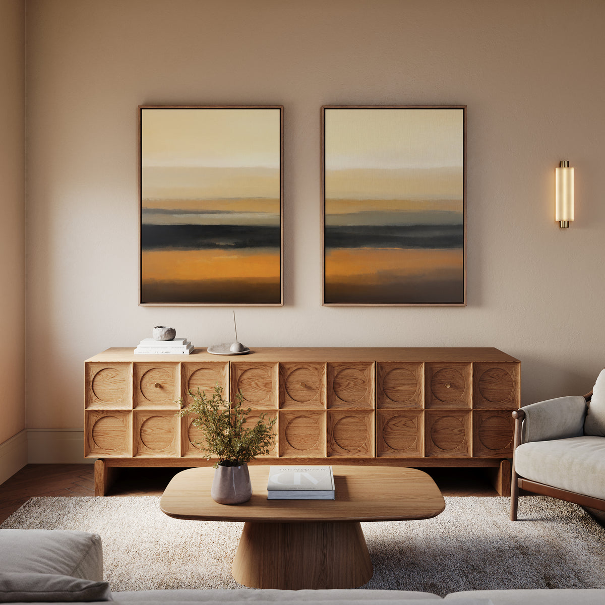 A modern living room featuring Desert Mirage - Set Of 2 abstract prints, a wooden sideboard, earth tone armchairs, a coffee table with a book and plant, and a wall light fixture.