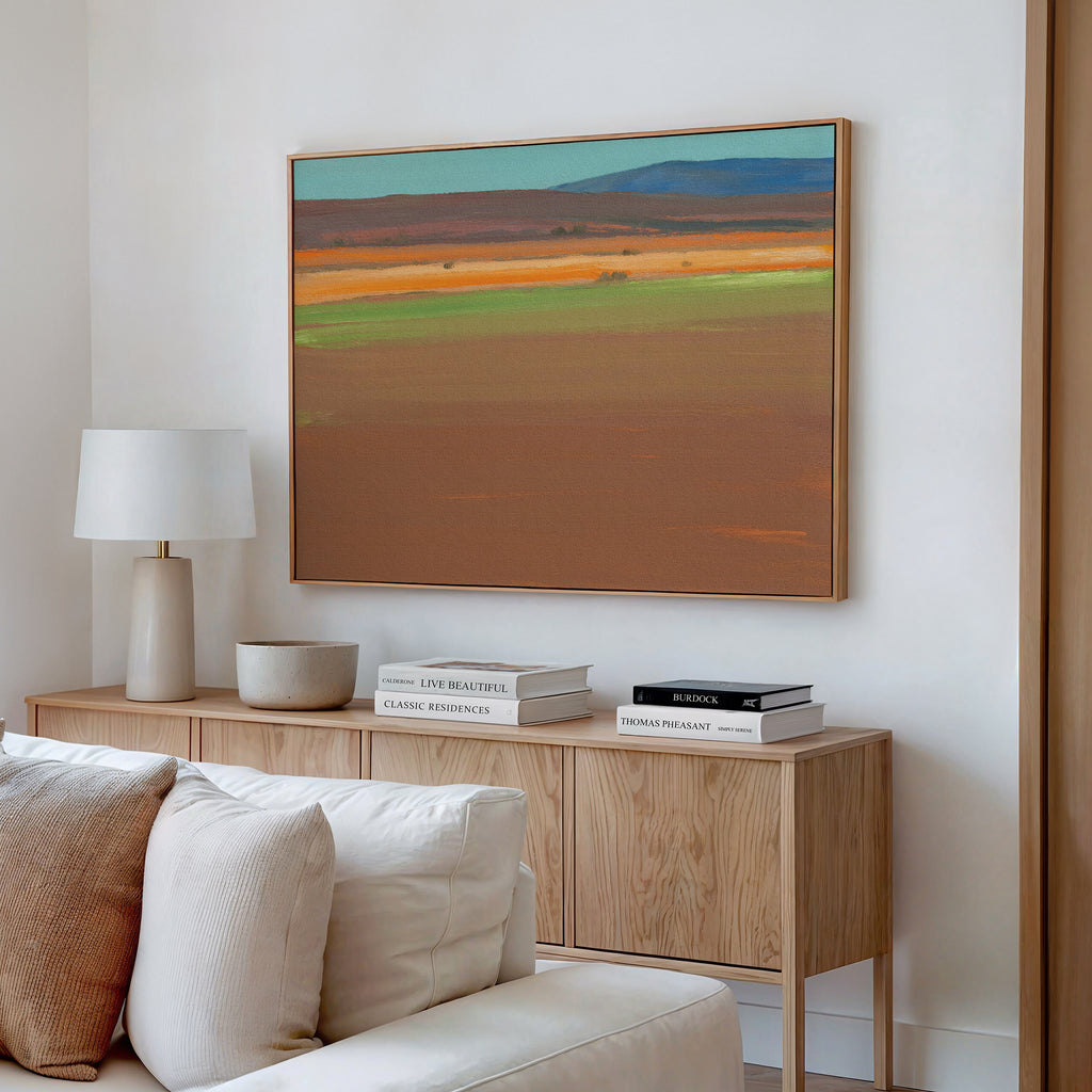 A modern living room with a light wood sideboard, books, a lamp, and a bowl is anchored by the Desert Plains Canvas Art - Horizontal above a cream sofa, bringing earthy and blue southwestern warmth to the white wall.