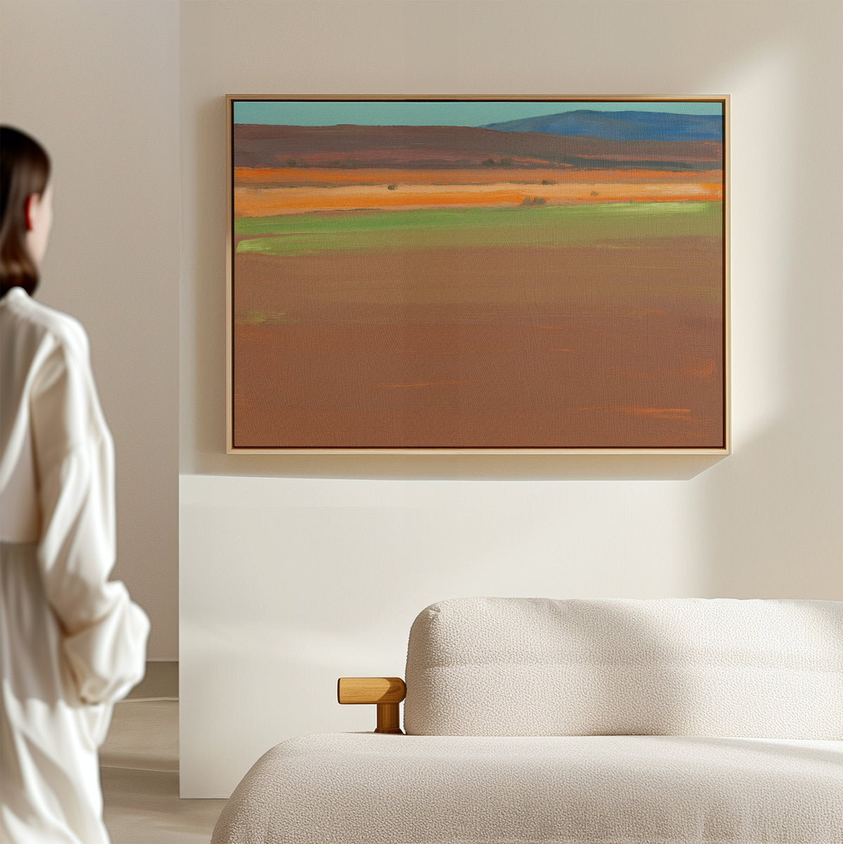 A person stands in a minimal, modern room with a beige sofa, admiring the "Desert Plains Canvas Art - Horizontal," an abstract landscape featuring earthy brown, orange, green, and blue tones that evoke southwestern warmth.