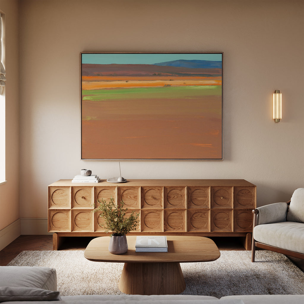 A modern living room with a wooden coffee table, light sofa, and textured sideboard exudes Southwestern warmth. The Desert Plains Canvas Art - Horizontal hangs above the sideboard, while a small vase of branches sits on the table.