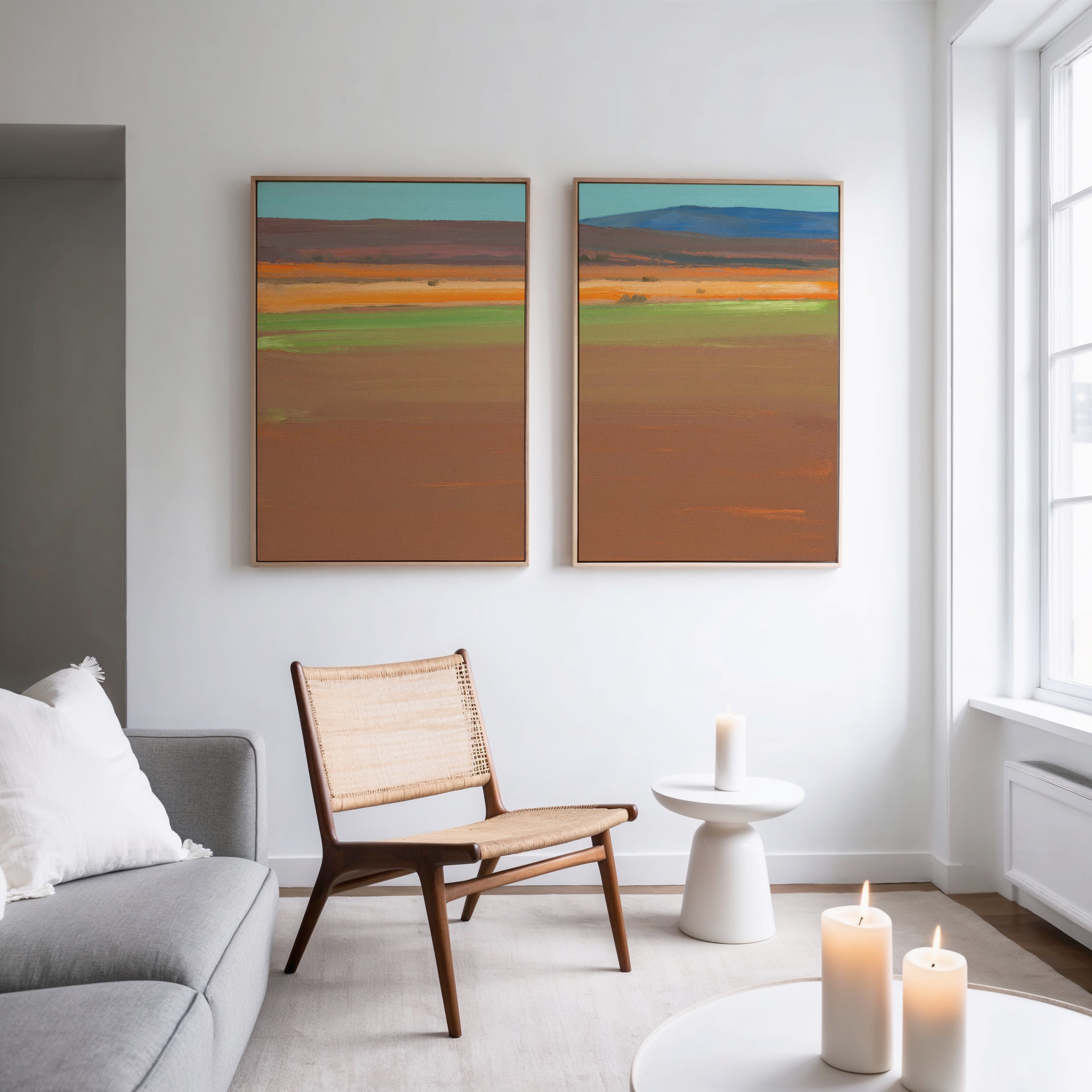 A modern living room with a gray sofa, wicker chair, and round white side table features three lit candles and the Desert Plains - Set of 2 colorful desert landscape canvas prints on a white wall. Natural light streams in from large windows.