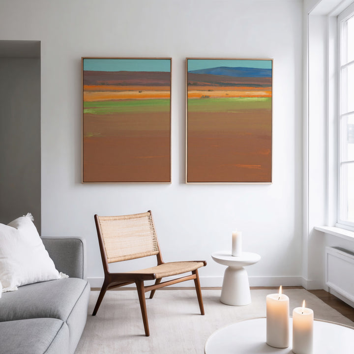 A modern living room with a gray sofa, wicker chair, and round white side table features three lit candles and the Desert Plains - Set of 2 colorful desert landscape canvas prints on a white wall. Natural light streams in from large windows.