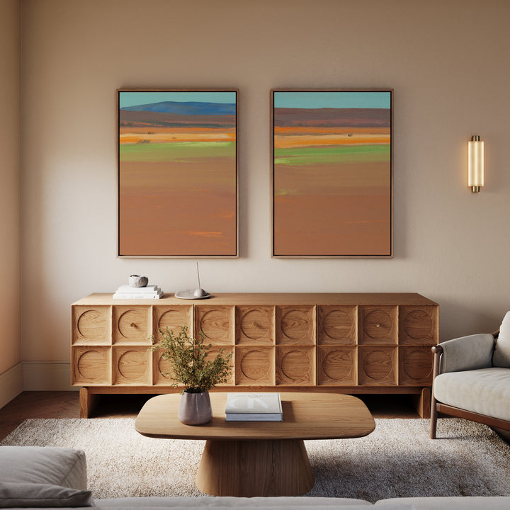 A modern living room featuring a wooden sideboard, the "Desert Plains - Set of 2" canvas prints above it, a wall light fixture, low wood coffee table with books, a vase of greenery, and a gray armchair on a rug.