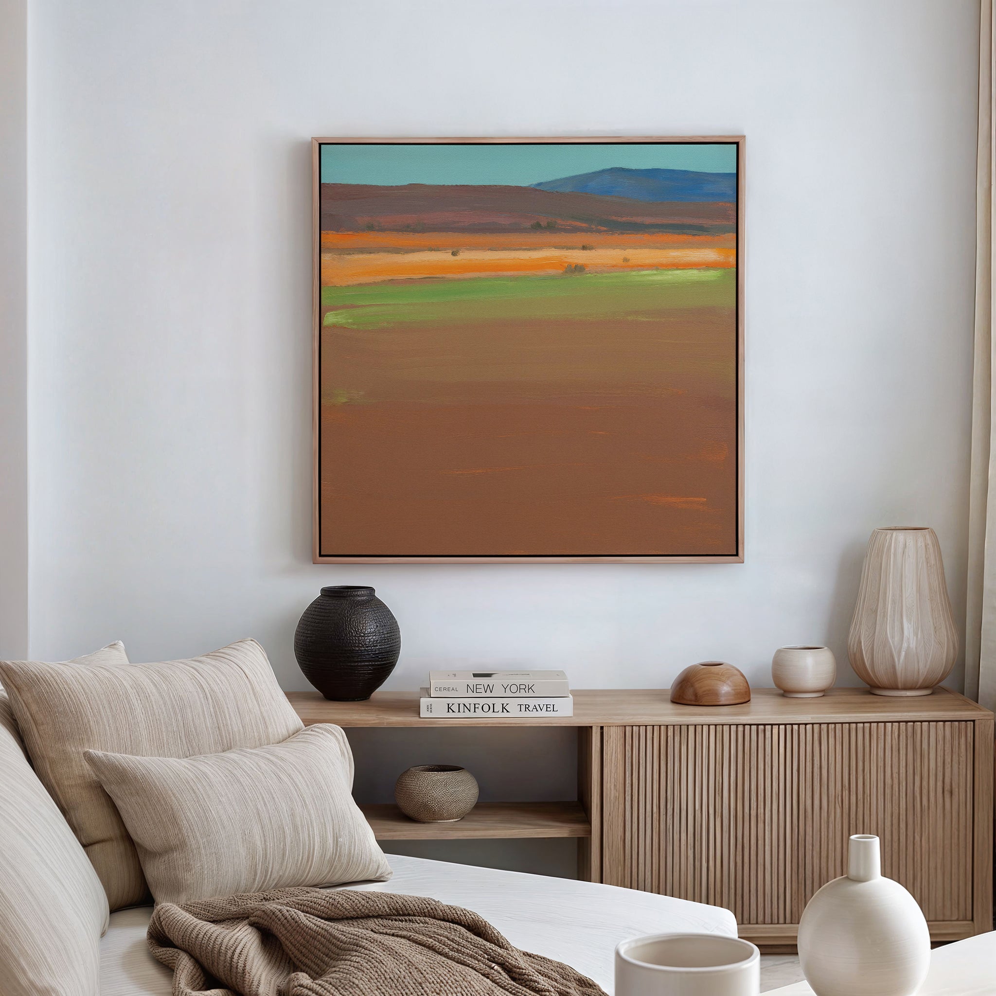 A minimal living room features a beige sofa with striped pillows, a wooden sideboard, and the Desert Plains Canvas Art - Square above as southwestern wall art, adding warmth and modern style to the neutral-toned space.